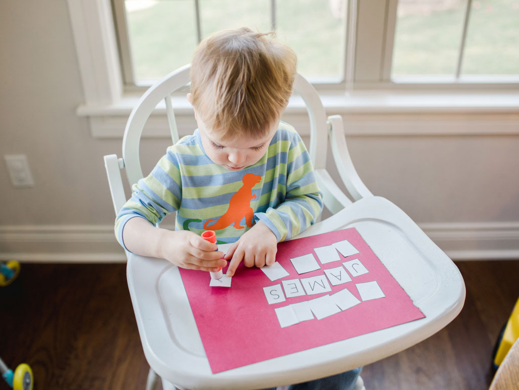 A boy in a highchair pasting paper letters onto a piece of construction paper