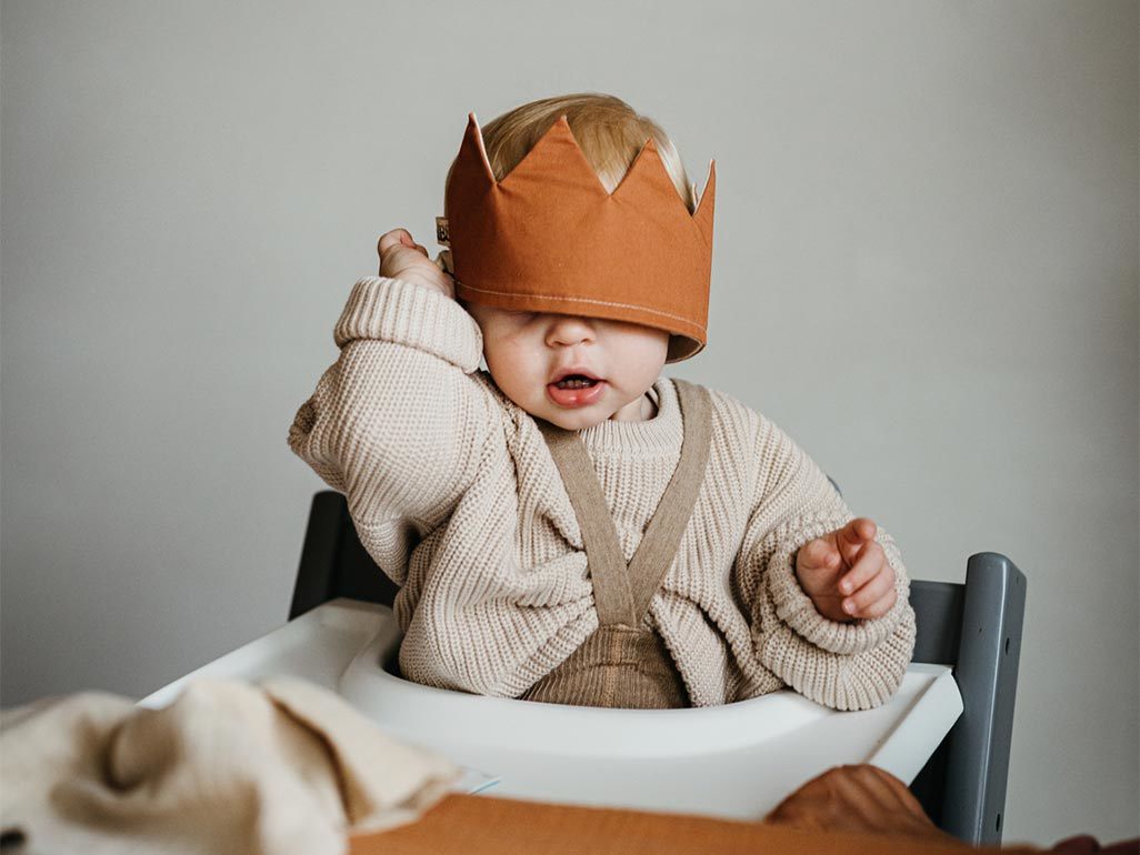 A baby in a highchair with a fabric crown covering their eyes